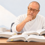多数の分厚いガイドを開き、英語版と日本語版を比較する野村隆昌